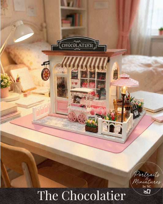 A miniature dollhouse model of a French chocolate shop called 'Chocolatier', displayed on a pink mat on a white desk in a cozy, softly-lit bedroom setting. The shop features a striped awning, detailed interior, and small accessories.