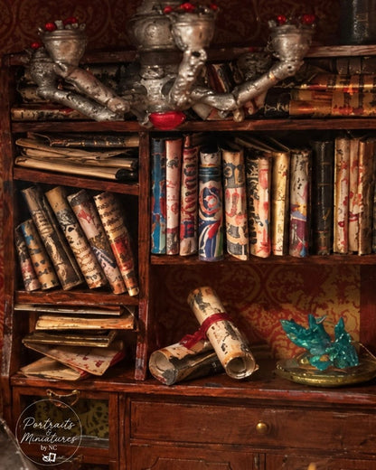 A miniature diorama bookshelf filled with aged scrolls, books, and decorative goblets, with a small blue crystal on a tray in the lower right corner.