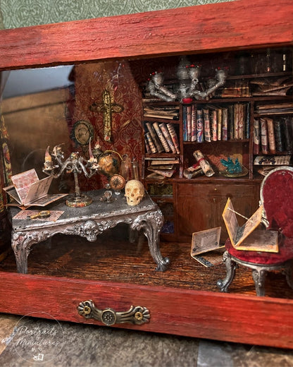 A miniature gothic study diorama inside a red wooden display case, featuring a detailed desk with a skull, candle holder, bookshelves, and a red upholstered chair.