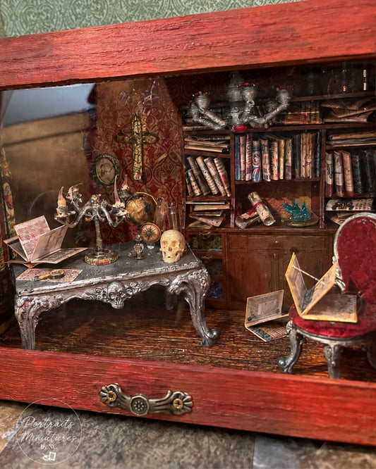 A miniature gothic study diorama inside a red wooden display case, featuring a detailed desk with a skull, candle holder, bookshelves, and a red upholstered chair.