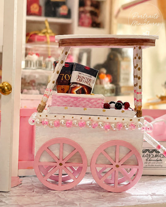 A decorative pink and white miniature cart or cart-like display stand with large pink wheels, pink and white striped supports, a light wood top, and pink floral trim, shown in a styled indoor setting with small props and a visible 'Portraits Memories' watermark.