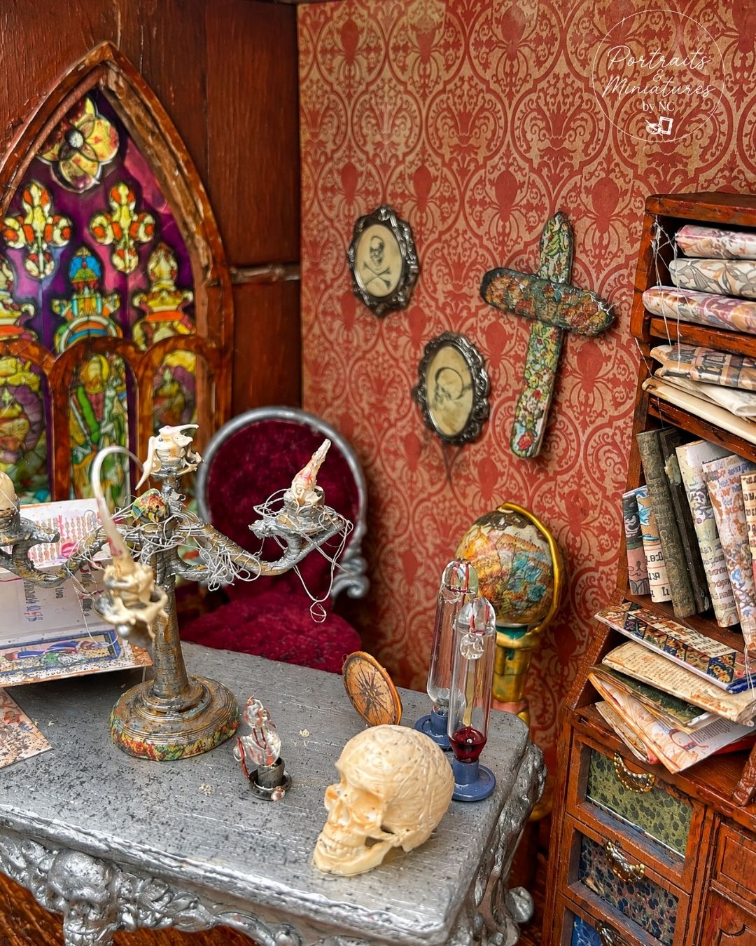 A miniature gothic-themed dollhouse interior featuring a detailed desk with a skull, ornate candle holder, miniature globe, framed portraits, a red velvet chair, and a bookshelf filled with tiny books and scrolls.