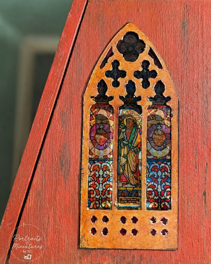 A close-up of a miniature decorative stained-glass window panel mounted on a red-painted wooden surface. The panel has an arched Gothic shape with three vertical window sections featuring colorful stained-glass-style illustrations and dark cutout details at the top and bottom.