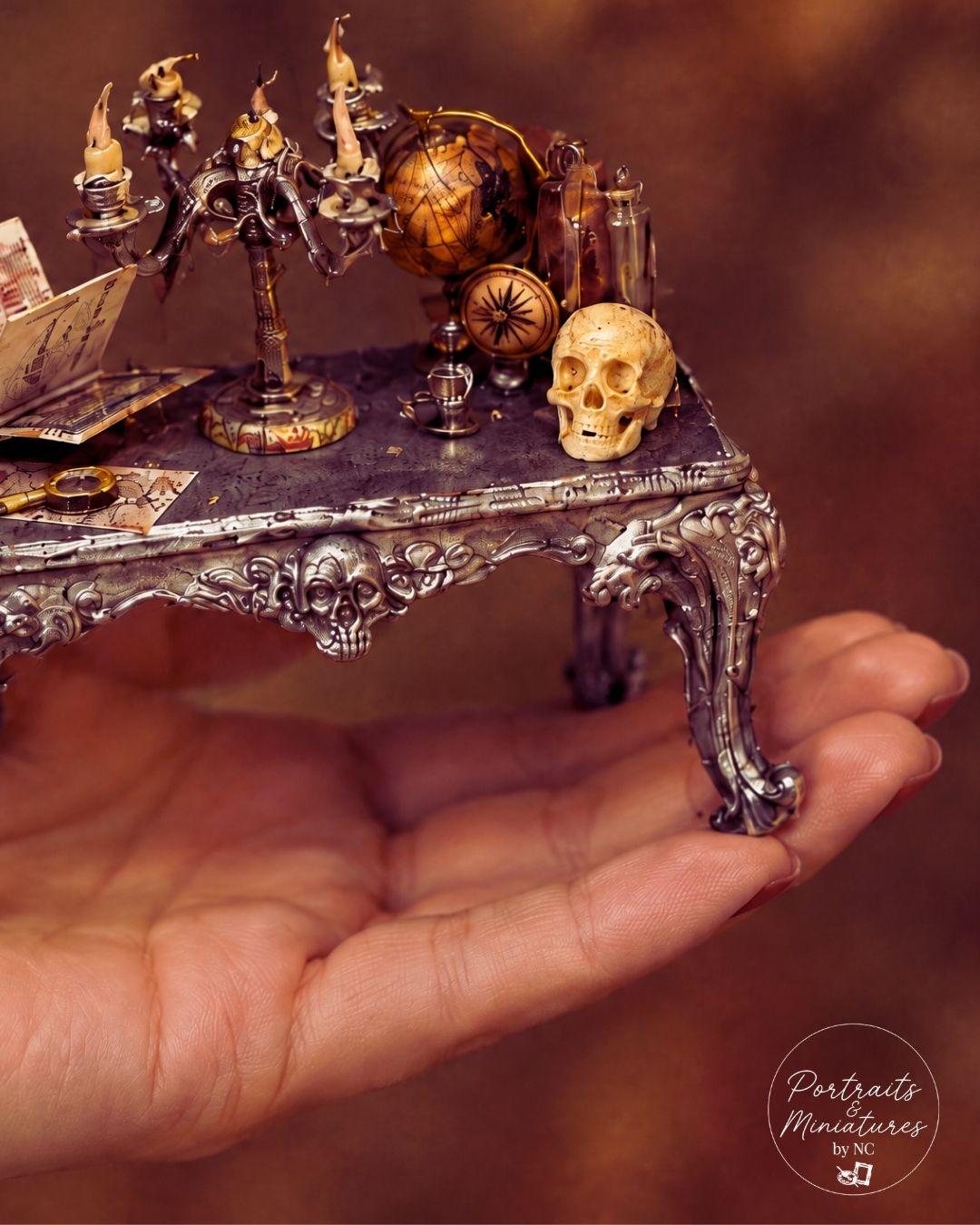 A miniature ornate silver and gold gothic table holding a skull, globe, and candlestick, shown resting on a person's hand against a warm brown background. A 'Portraits Miniatures by ND' logo is visible in the bottom right corner.