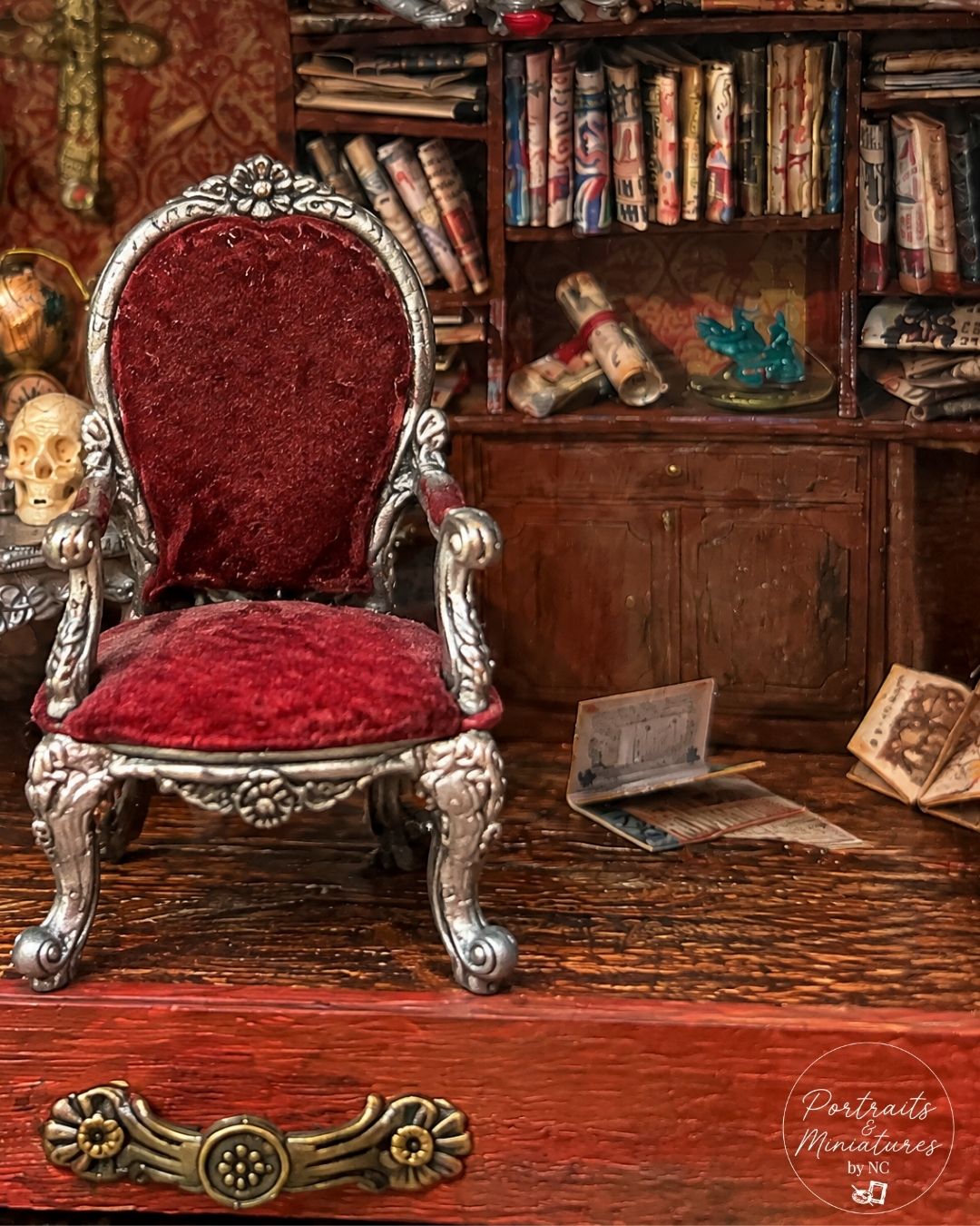 A miniature gothic-style armchair with ornate silver carved metal legs and a deep red velvet upholstered back and seat, set in a detailed miniature study with a bookshelf, skull, and old papers.