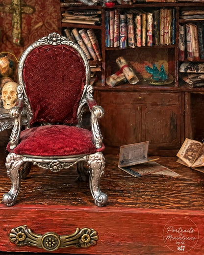 A miniature gothic-style armchair with ornate silver carved metal legs and a deep red velvet upholstered back and seat, set in a detailed miniature study with a bookshelf, skull, and old papers.