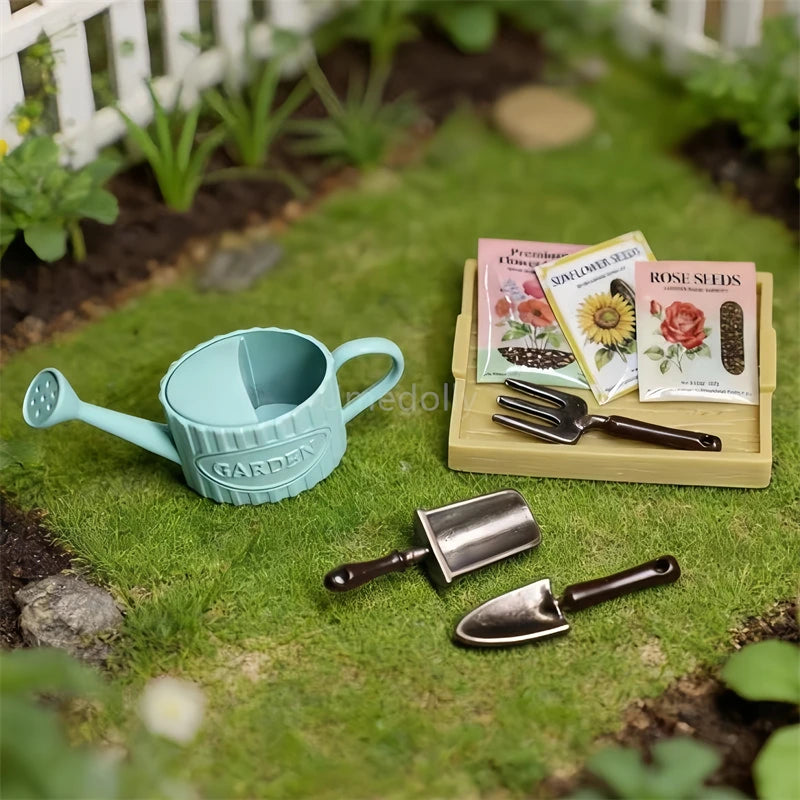 A miniature garden toy set arranged on artificial grass, including a light blue watering can, two small metal trowels, a wooden-handled garden fork, and three seed packet cards labeled Vegetable Vines, Sunflower Seeds, and Rose Seeds.