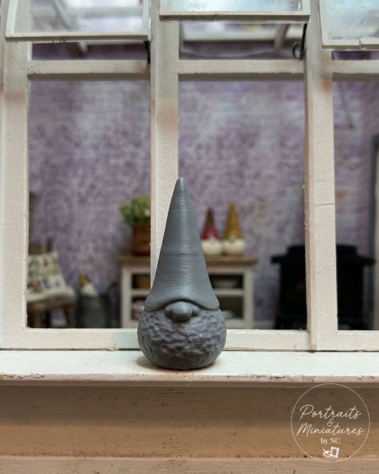 A small gray gnome 1/12 scale  figurine with a textured beard and pointed hat sits on a white window sill, looking out through the window panes into a softly blurred room with lavender walls.