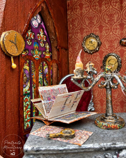 A miniature decorative scene featuring a small open book with printed pages, a silver candelabra with a lit candle, a round clock, and framed portraits on a tabletop against ornate red patterned wallpaper and a stained-glass-style window.