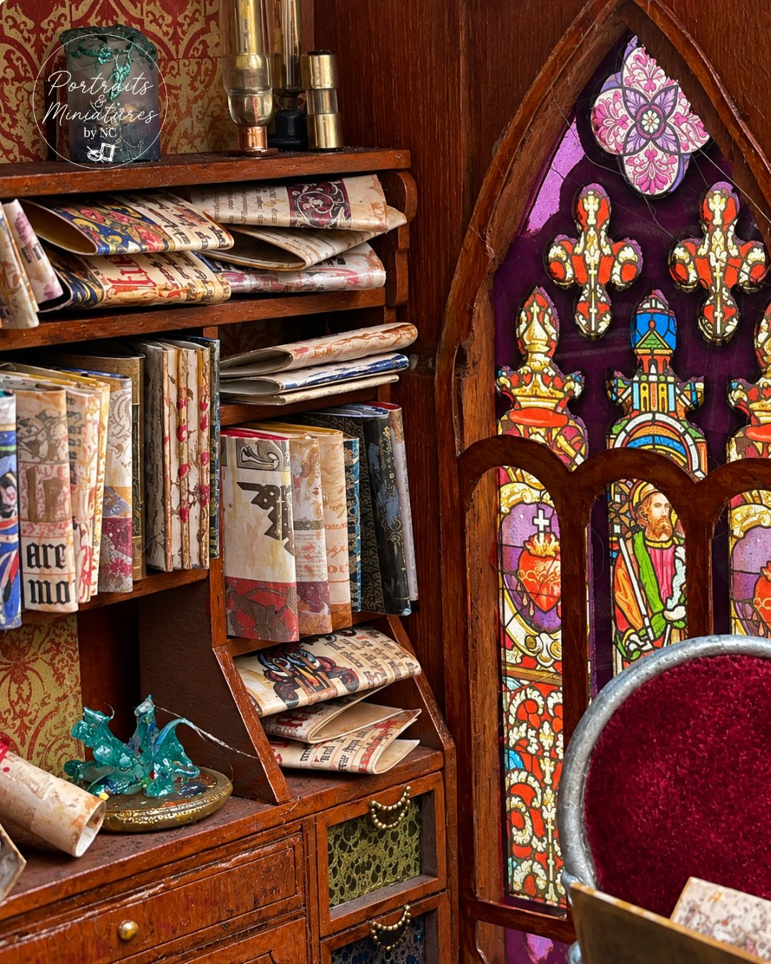A miniature diorama room scene featuring a detailed wooden bookshelf filled with miniature books, scrolls, and decorative items, next to a stained-glass window and a red velvet chair.
